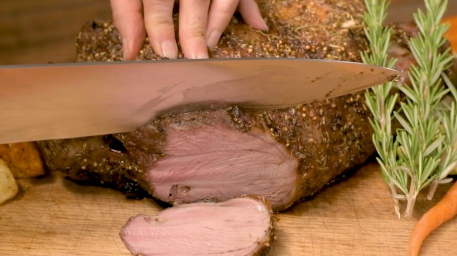 Cooked leg of lamb being sliced on a wooden board, showing juicy pink interior, with rosemary and carrots on the side.