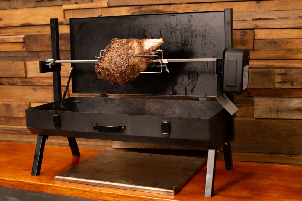 Leg of lamb cooking on a Flaming Coals Mini Spit rotisserie with motorised skewer, set up on a wooden bench against a timber wall.