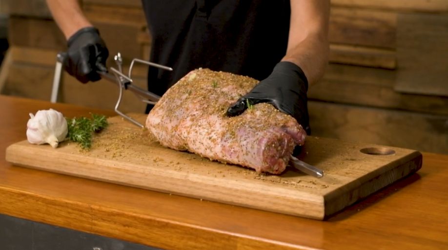 Seasoned leg of lamb being secured onto a rotisserie skewer with prongs, placed on a wooden board with garlic and rosemary beside it.