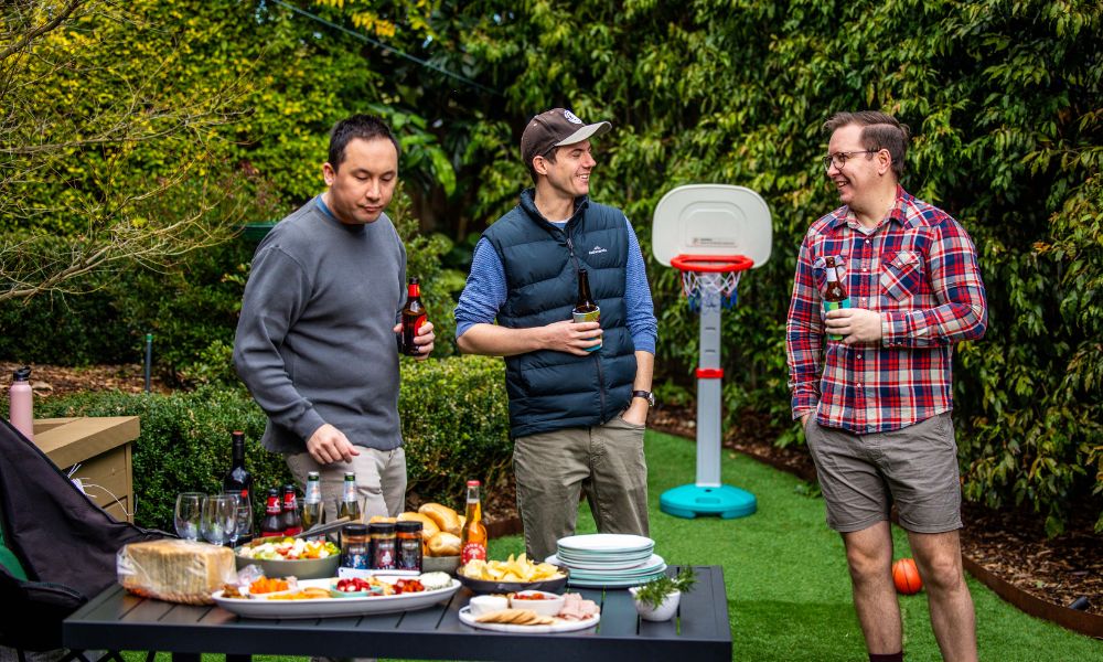 Three friends enjoy drinks and conversation outdoors around a table set with snacks, condiments, and drinks. They stand on a backyard lawn near a small basketball hoop, creating a relaxed and friendly BBQ atmosphere.