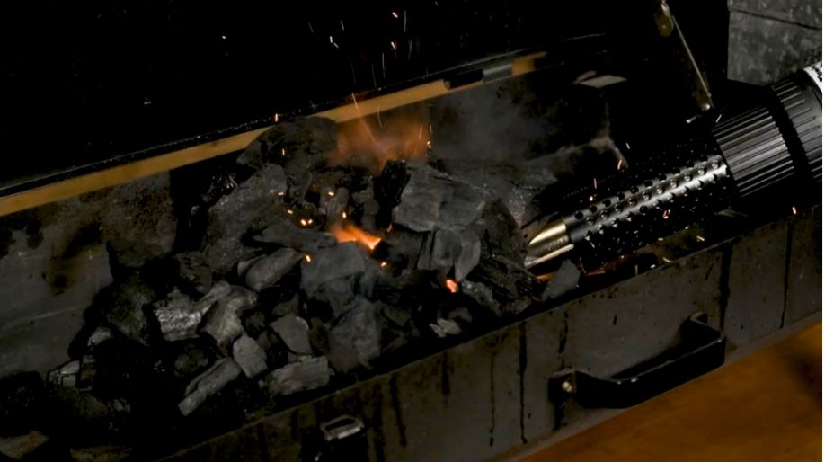 Charcoal being lit inside a spit roaster using an electric charcoal starter wand, with sparks and flames visible among the coals.