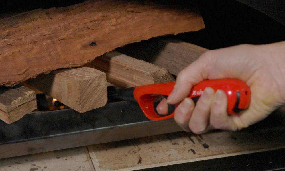 A Fire lighter being used to ignite kindling wood inside a wood-fired pizza oven, with larger firewood stacked above the kindling.