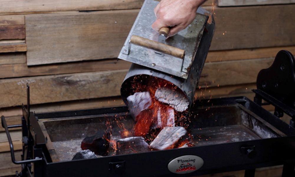 Hand pouring lit charcoal from a chimney starter into a Flaming Coals Cyprus spit roaster, with glowing embers and sparks flying.
