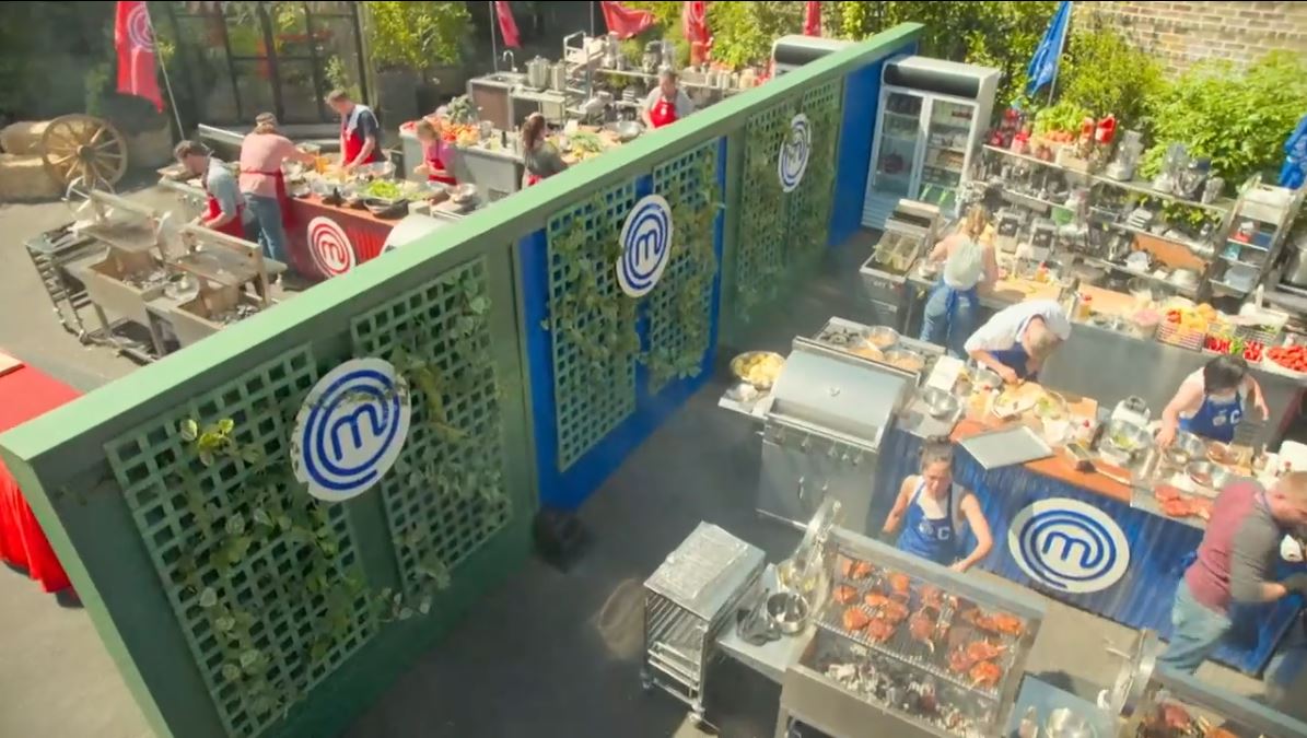 Aerial view of a MasterChef USA outdoor cooking challenge, with contestants cooking on Parrilla grills, preparing dishes in a busy kitchen setup surrounded by greenery and MasterChef branding.