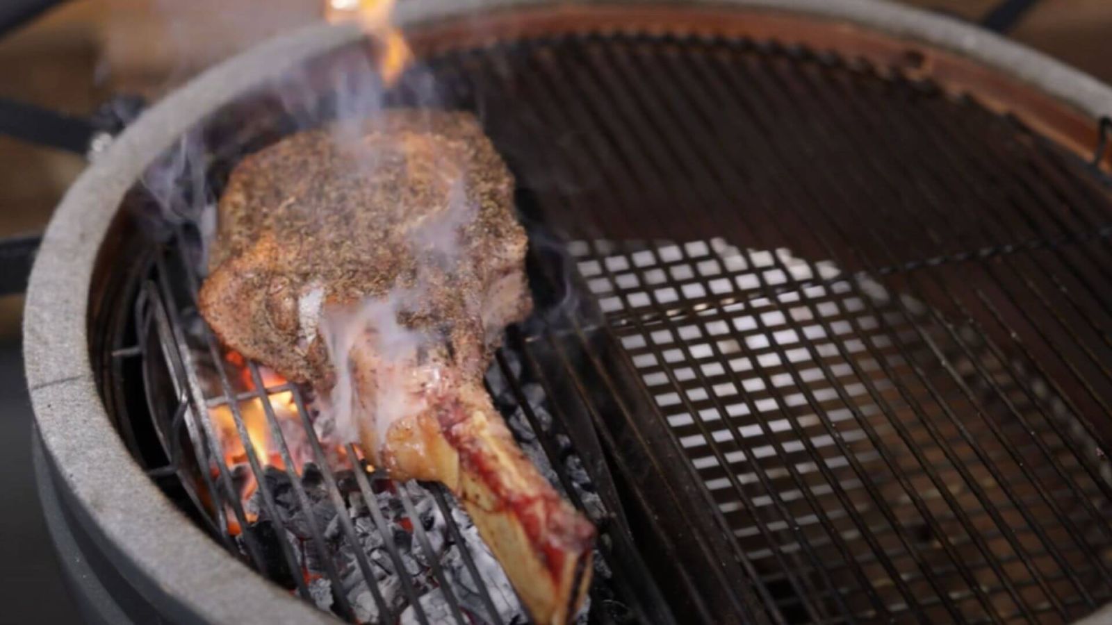 Tomahawk steak searing over charcoal in a ceramic Kamado smoker, with smoke rising for authentic BBQ flavour.