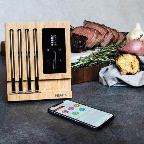 A Meater Block with four probes is displayed next to a smartphone showing the Meater app. The setup is placed on a countertop with perfectly cooked slices of beef, garlic, and herbs in the background.