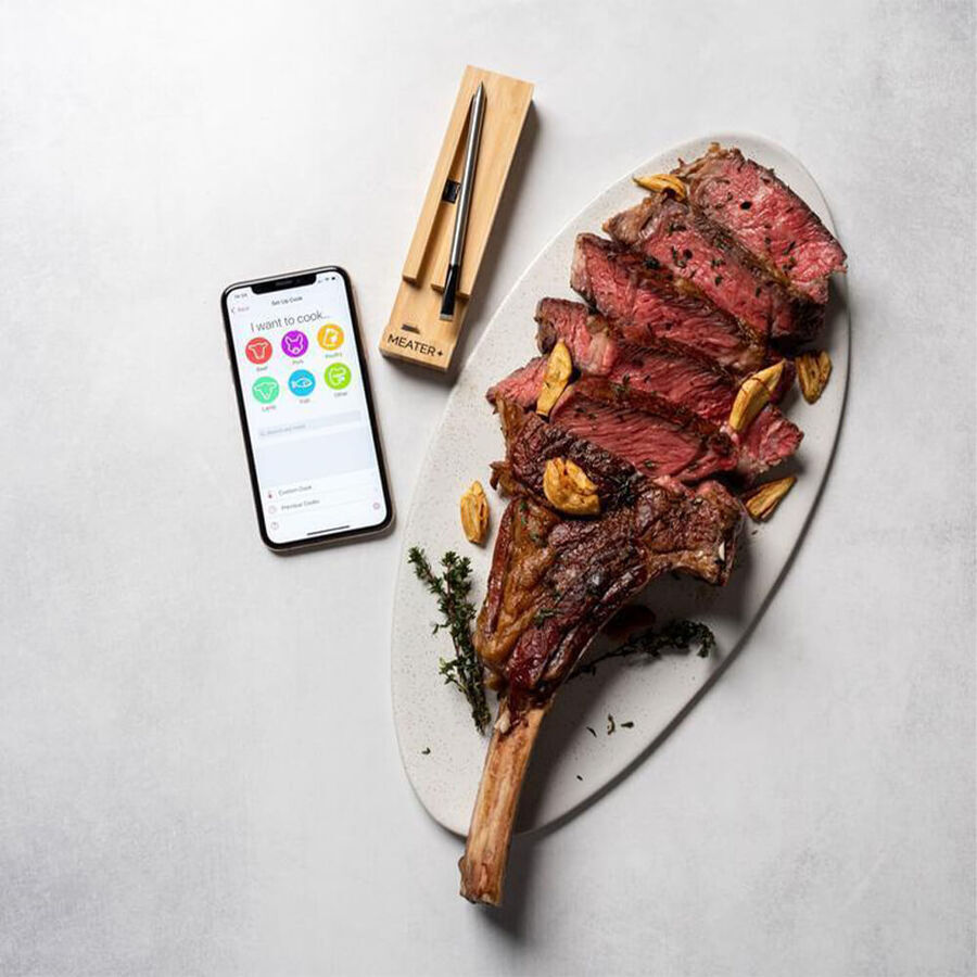 A beautifully cooked steak sliced and served on a white platter with garlic and herbs, next to a Meater Plus thermometer resting on a wooden stand. A smartphone with the Meater app open is also visible beside the meal.
