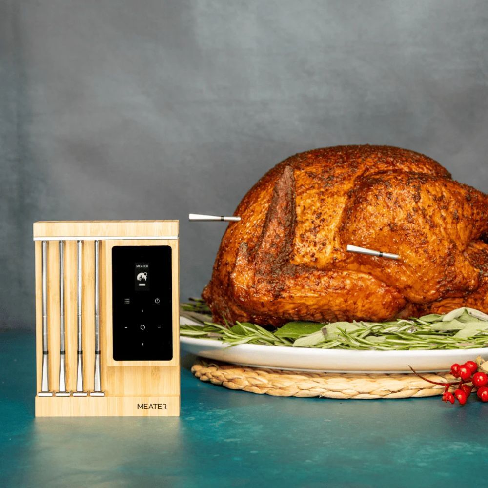 A perfectly roasted turkey placed on a serving platter with fresh herbs, featuring two Meater Pro thermometer probes inserted into the bird. The Meater Pro unit is positioned beside the turkey, showing its sleek wooden design and digital display.