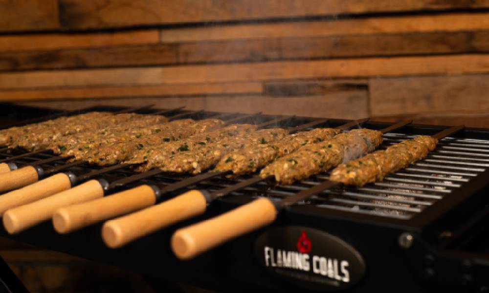 Raw minced meat kebabs cooking on flat skewers over a Flaming Coals Cyprus Spit grill, with smoke rising as they begin to sizzle.