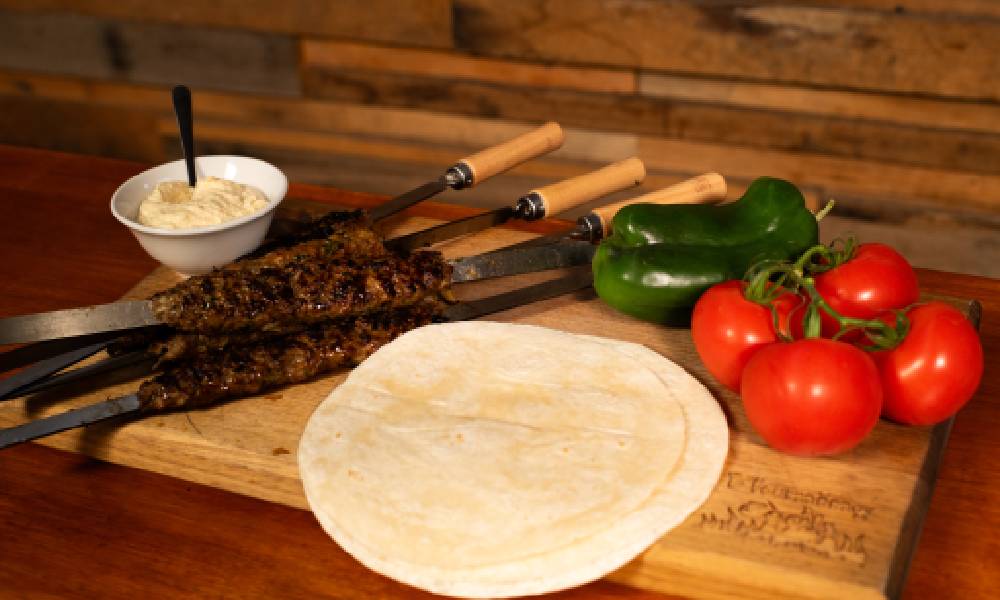 Grilled minced meat kebabs on flat skewers served on a wooden board, accompanied by flatbread 