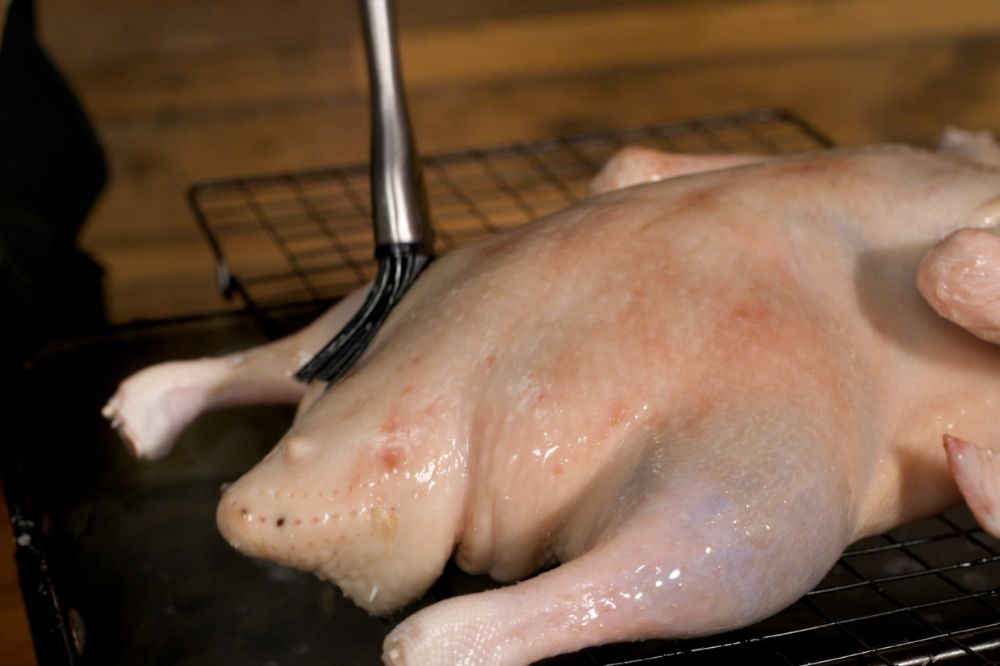 Whole raw duck on a roasting rack being brushed with a honey and hot water mixture to prepare the skin for spit roasting.