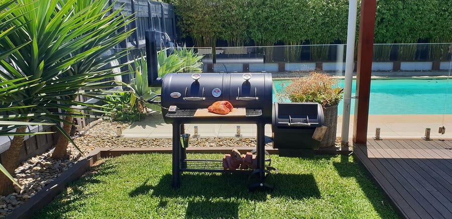 Offset smoker in a backyard by the pool with a piece of meat resting on the cooking surface.