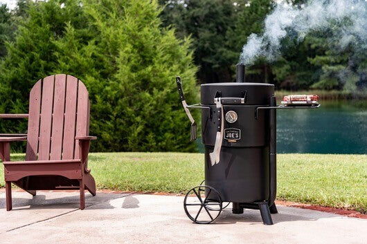 Drum smoker with smoke rising, set on a patio next to a wooden chair and a scenic lake.