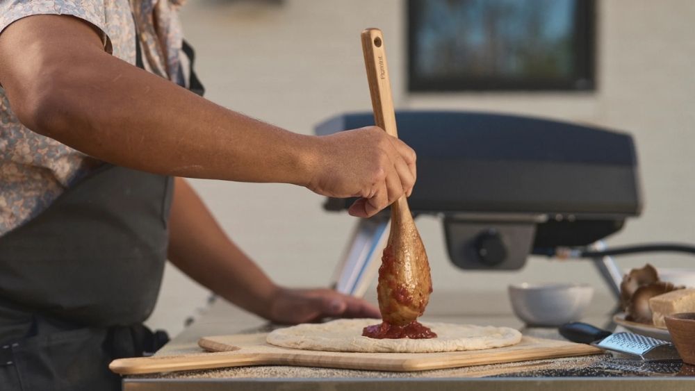 A person spreading tomato sauce on pizza dough using a wooden spoon, with an Ooni Koda 2 pizza oven in the background."