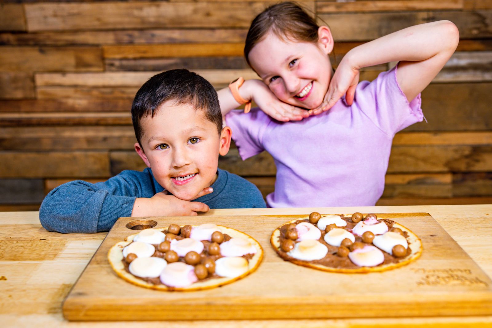 Two smiling children posing with homemade dessert pizzas topped with marshmallows and chocolate balls.