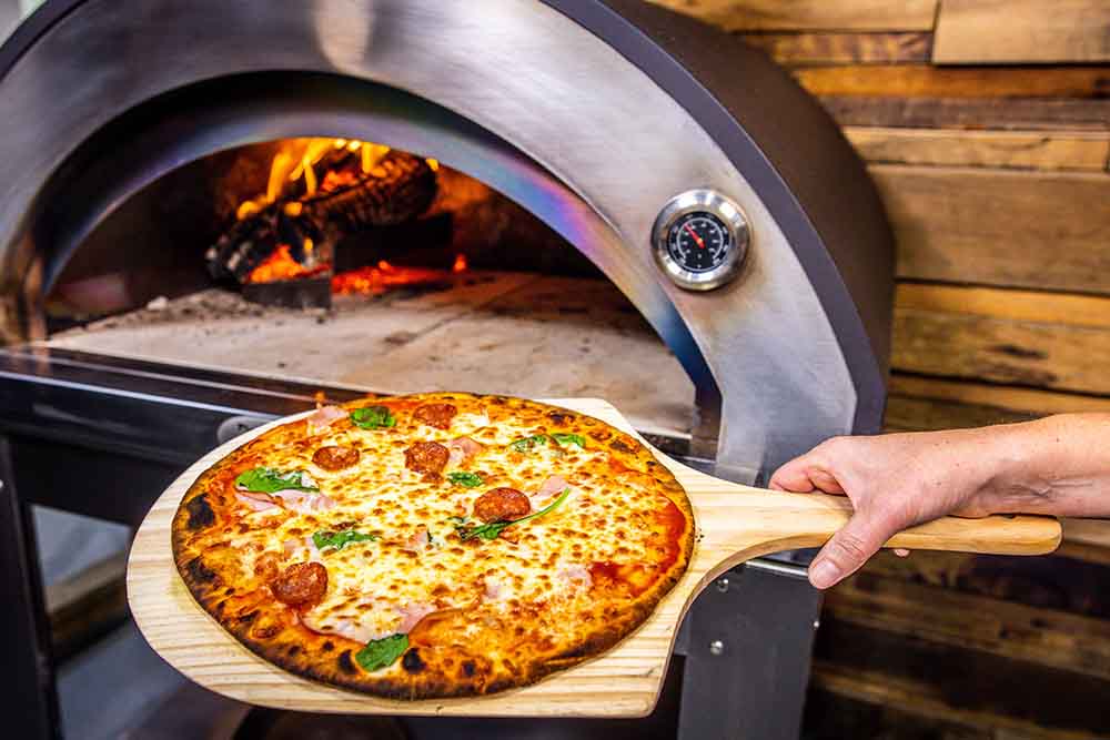 Wood-fired pizza oven with a freshly baked pizza on a wooden peel, showing a golden crust with melted cheese and toppings, and flames visible inside the stone oven.