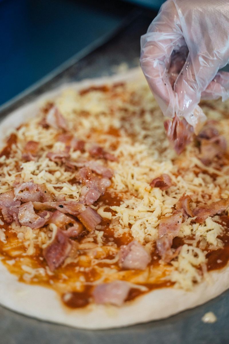 Pizza base topped with tomato sauce, shredded cheese, and leftover BBQ meat pieces being added by hand with a glove. Photo by pexels-anntarazevich-6937416
