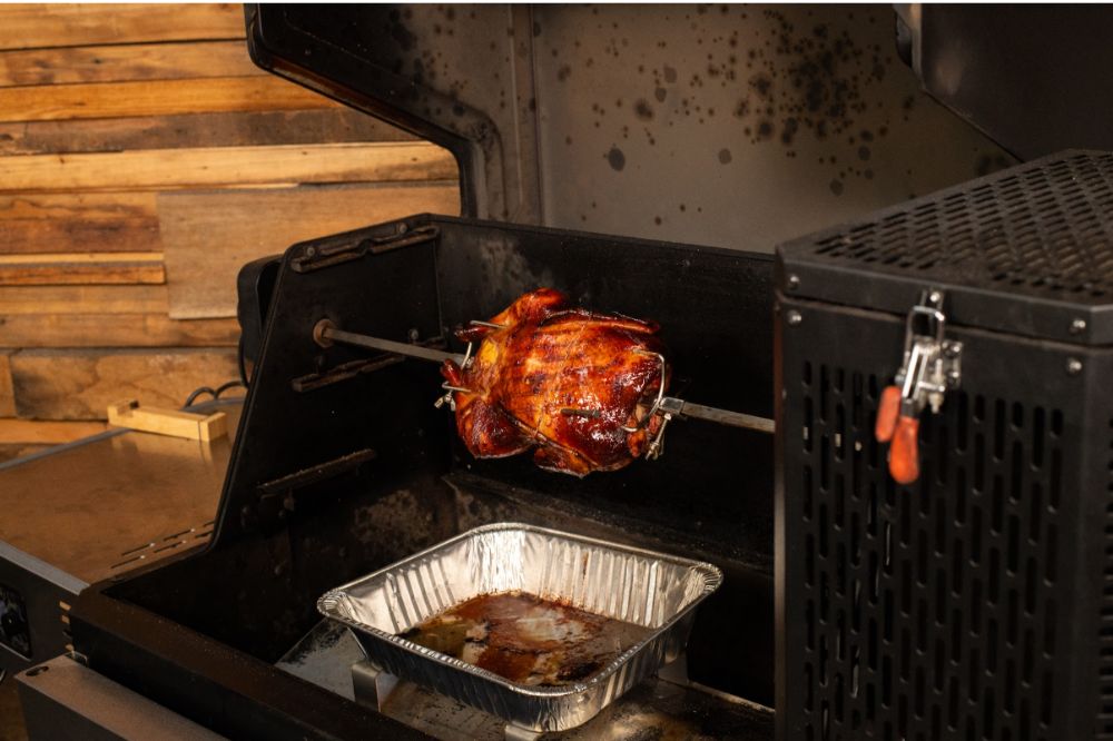 Whole duck cooking on a spit rotisserie inside a Masterbuilt Smoker, with a drip tray positioned underneath to catch juices.