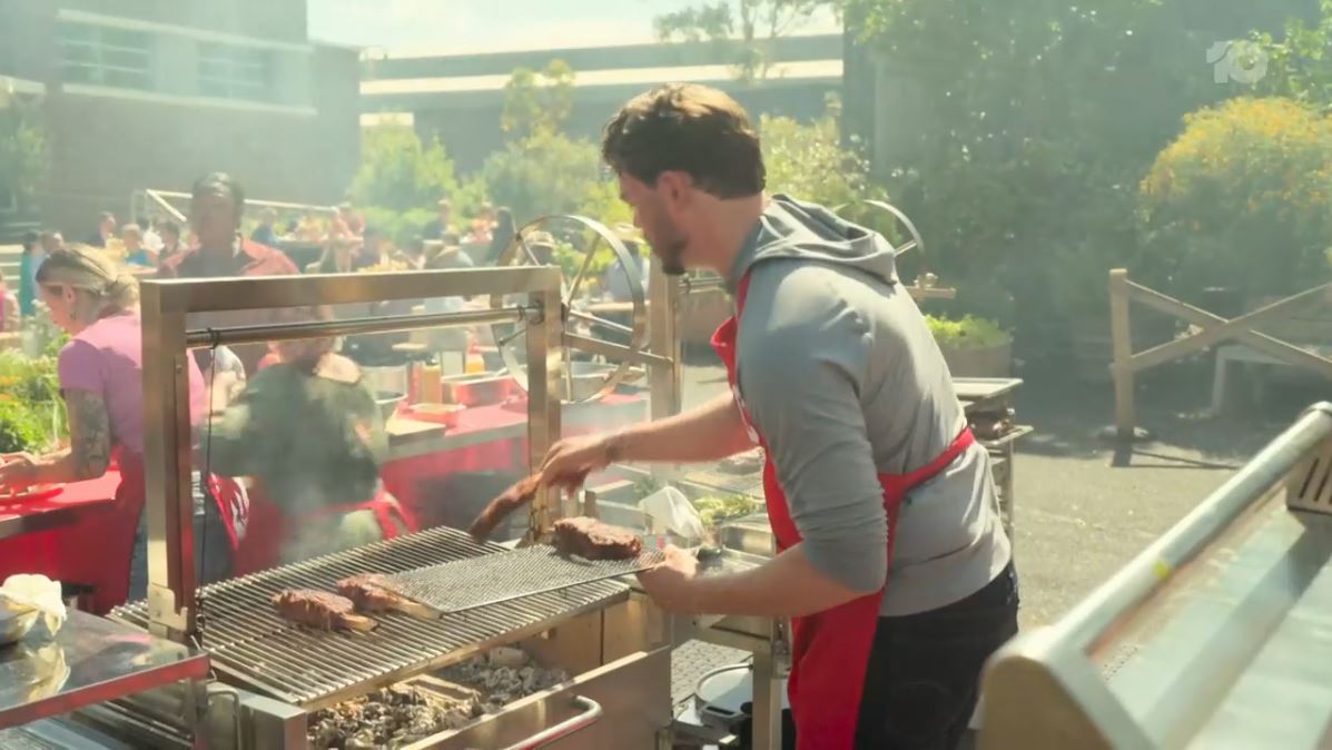 Contestant placing meat onto a Parrilla grill, with a smoky outdoor cooking environment and other participants working in the background during a MasterChef challenge.