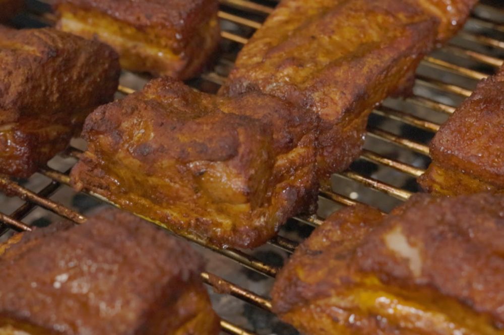 Seasoned pork belly bites cooking on a wire rack inside an SNS Kamado grill. The pork is developing a smoky bark with a rich, caramelized crust during the low-and-slow smoking process.          Ask ChatGPT