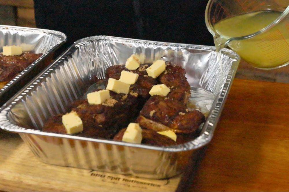 Smoked pork belly bites placed in a foil tray, each topped with cubes of butter and a sprinkle of brown sugar. 