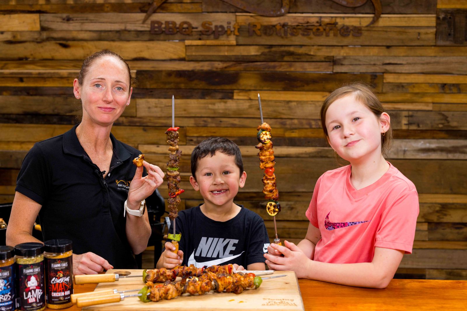 Family BBQ fun with kids holding skewers of grilled chicken, lamb, and pork, seasoned with Flaming Coals rubs, showcasing outdoor cooking and rotisserie skewer recipes.