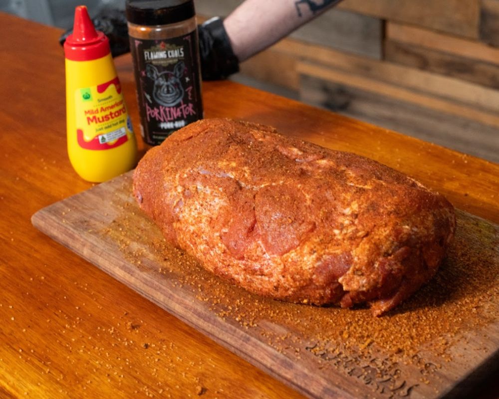 Pork shoulder coated with Flaming Coals Porkinator rub and mustard on a wooden board ready for smoking.