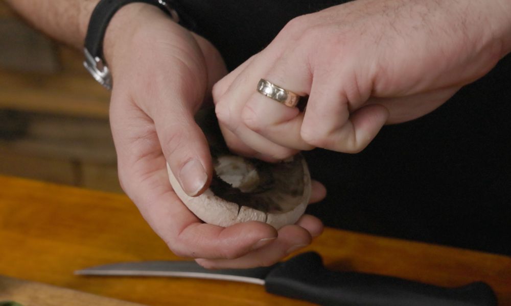 Hands removing the stem from a raw mushroom cap during preparation, with a knife resting on a wooden board nearby.
