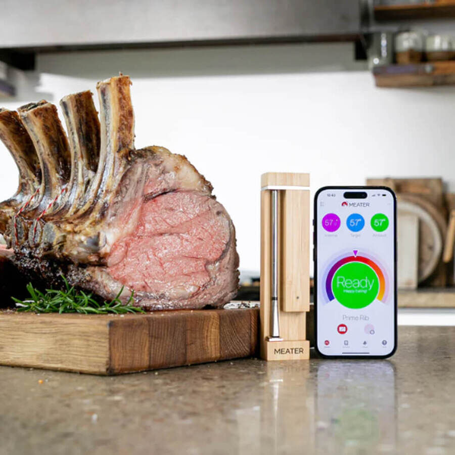 A perfectly cooked prime rib roast rests on a wooden board beside the Meater Pro Wireless Meat Thermometer and a smartphone displaying the Meater app. The app screen shows the internal temperature and readiness status, demonstrating precise wireless cooking control.
