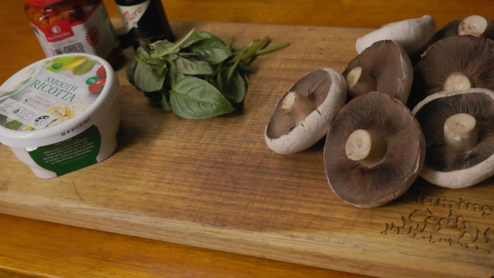 Ingredients for stuffed mushrooms laid out on a wooden board, including whole raw mushrooms, fresh basil, smooth ricotta, sun-dried tomatoes, and balsamic glaze.