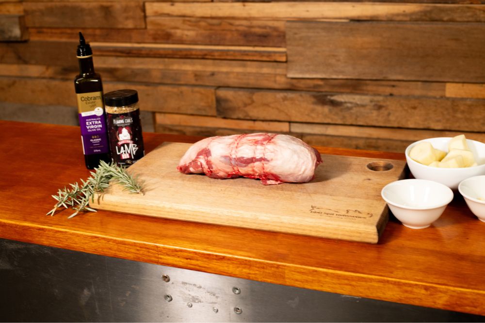 Raw boneless lamb roast on a wooden cutting board, surrounded by rosemary, Flaming Coals Lamb Rub, Cobram Estate extra virgin olive oil, and bowls of chopped potatoes and seasoning ingredients, ready for preparation.