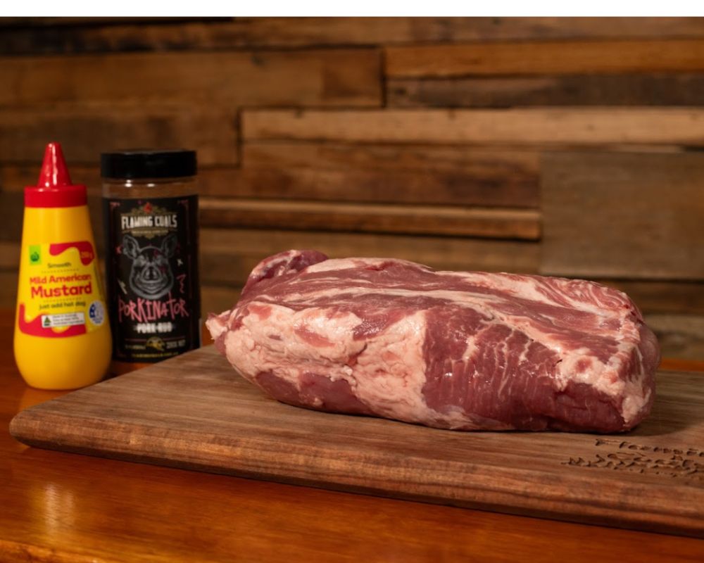 Raw pork shoulder on a wooden board with Flaming Coals Porkinator rub and mustard ready for Seasoning. 
