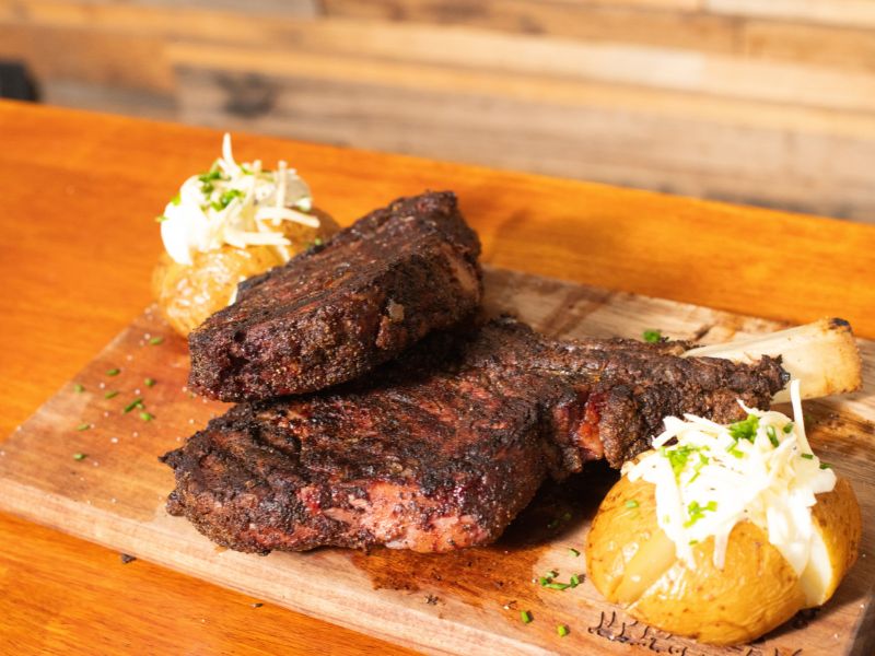 Reverse sear tomahawk steaks served with baked potatoes topped with sour cream, cheese and chives on a wooden board.
