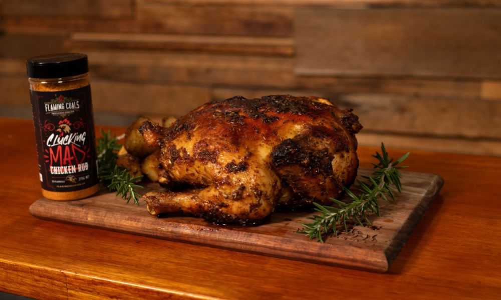 A perfectly roasted whole chicken seasoned with Flaming Coals Clucking Mad Chicken Rub, resting on a wooden cutting board garnished with fresh rosemary