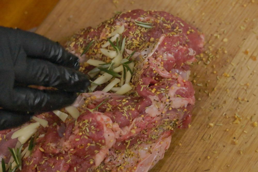 Close-up of a gloved hand stuffing fresh rosemary and sliced garlic into a seasoned lamb roast, ready for tying and cooking.