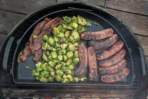 Slow ’N Sear Cast Iron Drip ’N Griddle Pan on a kettle grill, cooking sausages, bacon, and Brussels sprouts over charcoal heat.