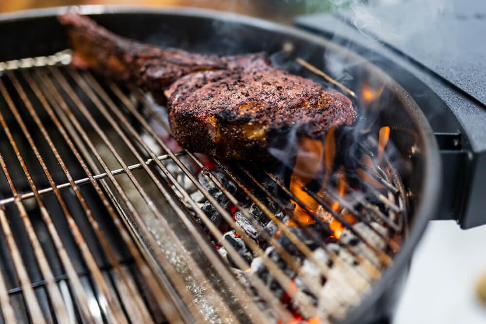 Close-up of a perfectly seared steak cooking over charcoal on an SNS Kettle BBQ, showing flames and smoke rising for that authentic grill flavour.