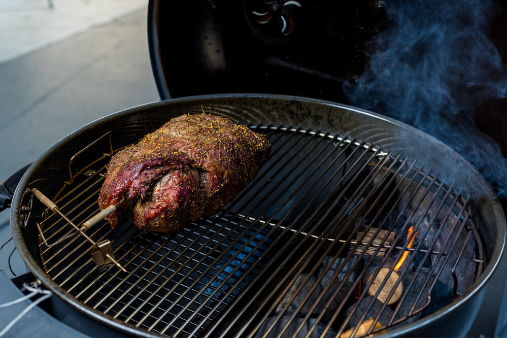 Herb-crusted beef roast cooking on a rotisserie inside an SNS Kettle BBQ, with charcoal glowing beneath for even heat and smoky flavour.