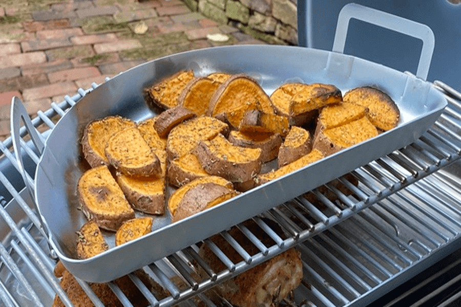 Slow N Sear Kettle Mini Drip Pan with handles, placed on a grill rack, holding sliced seasoned sweet potatoes.