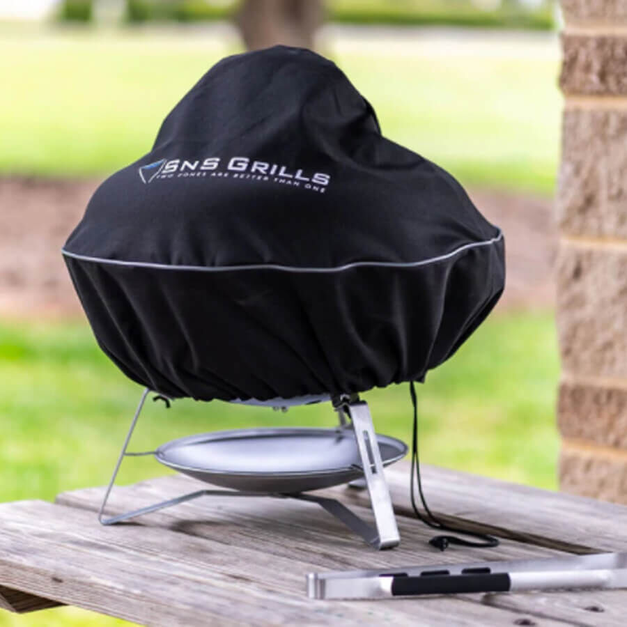 SNS Grills Travel Kettle Grill covered with a black protective cover, sitting on an outdoor wooden table with BBQ tongs nearby.