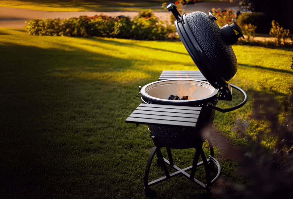 Ceramic Kamado smoker with open lid and charcoal fire burning inside, set up outdoors on a grassy lawn with foldable side shelves.