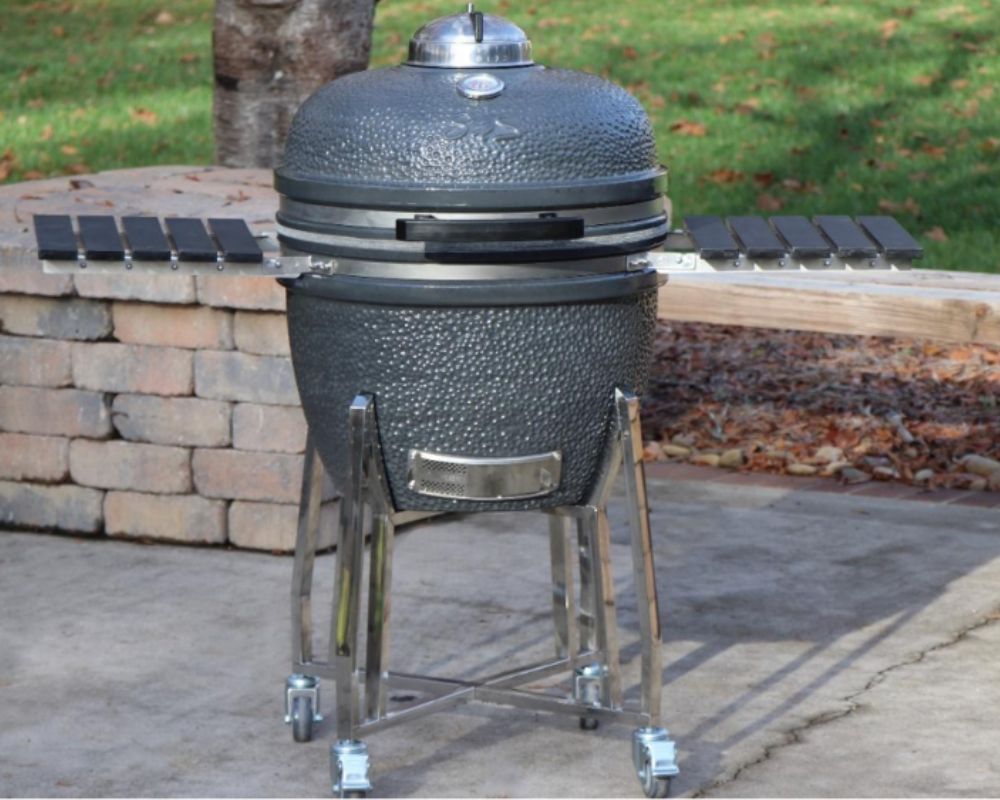 An SNS Kamado grill with a robust black ceramic body, equipped with a stainless steel base and adjustable vents for temperature control. It features foldable side shelves and sits on sturdy wheels, making it easy to move in an outdoor setting.