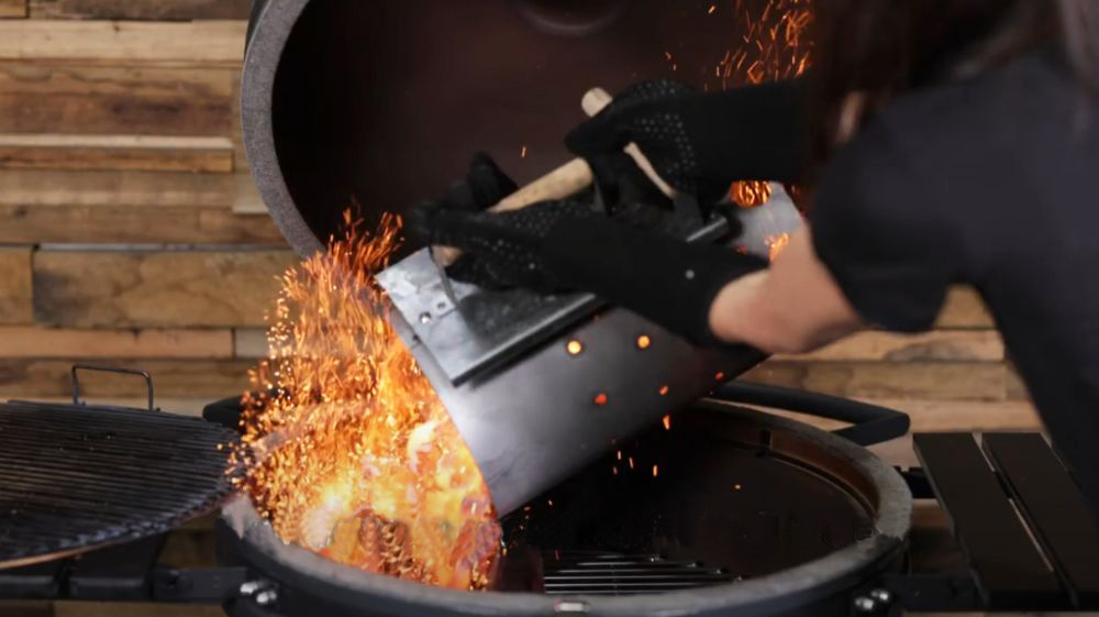 Person pouring lit charcoal from a chimney starter into an SNS Kamado grill, with sparks flying, preparing for a high-heat cook.