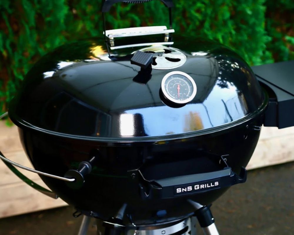 Close-up of an SNS Grills kettle BBQ with the lid closed, featuring an integrated temperature gauge and adjustable air vent.