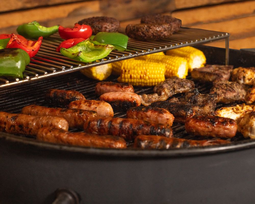Close-up of a Slow ‘N Sear Kettle BBQ grilling sausages, chicken wings, lamb chops, corn on the cob, burger patties, and capsicum for a mixed barbecue feast.