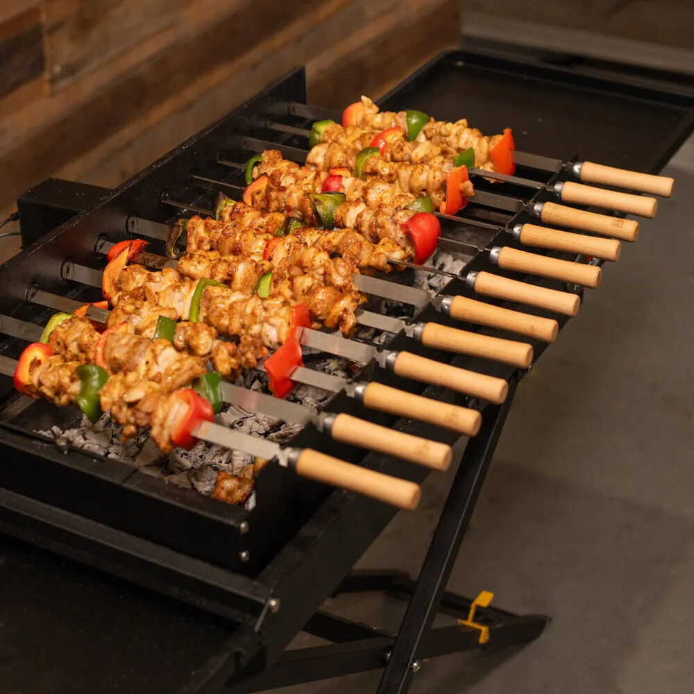 Chicken and vegetable skewers cooking over charcoal on a portable shashlik grill with wooden handled flat skewers.