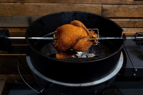 A whole seasoned chicken roasting over charcoal on a Flaming Coals 57cm Kettle Rotisserie Kit. The rotisserie rod spins evenly above the coals inside a black kettle BBQ, ensuring juicy, perfectly cooked results. A whole seasoned chicken roasting over charcoal on a Flaming Coals 57cm Kettle Rotisserie Kit. The rotisserie rod spins evenly above the coals inside a black kettle BBQ, ensuring juicy, perfectly cooked results.