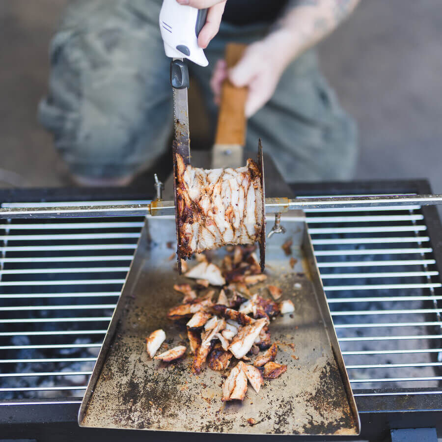 Stainless steel gyros carving shovel catching sliced meat while carving from a rotating gyros spit over a grill