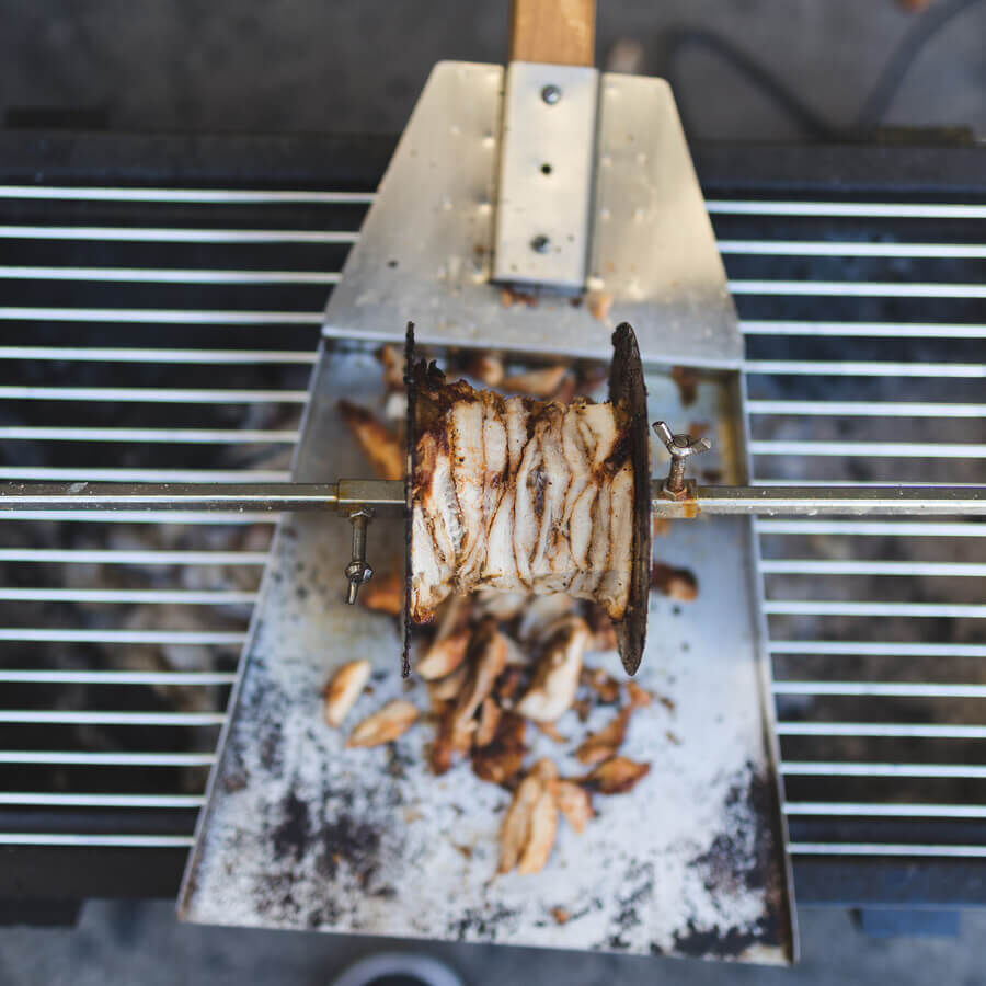 Stainless steel gyros carving shovel positioned beneath a rotating gyros spit collecting freshly carved meat over a charcoal grill