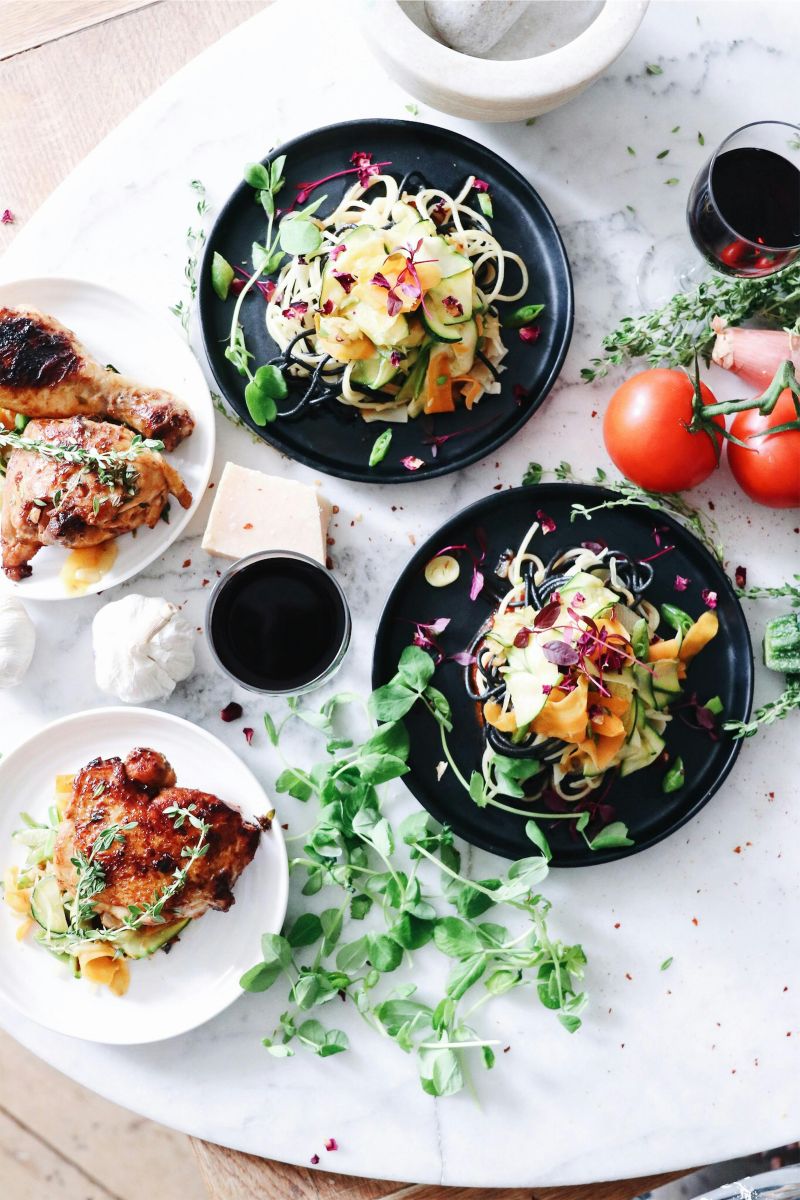 Colourful salads with zucchini noodles, fresh herbs, tomatoes, and grilled chicken served on black and white plates on a marble table.